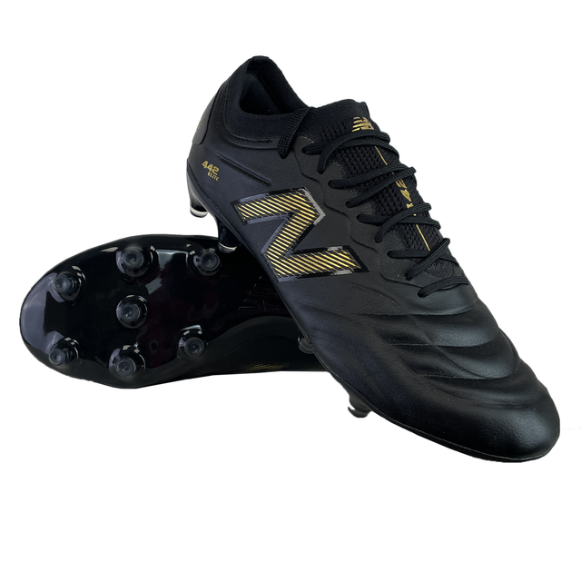 The New Balance 442 v2 Elite FG Wide Boots in Black/Metallic Gold feature a pair with one upright and the other on its side to display the studded sole, perfect for those needing wide boots for firm ground.