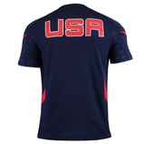 The USA Rugby 25/26 Training Poly Shirt by Macron is navy blue with "USA" in bold red and white on the back, red accents on the sides and sleeves, and made from breathable Poly Dry fabric.