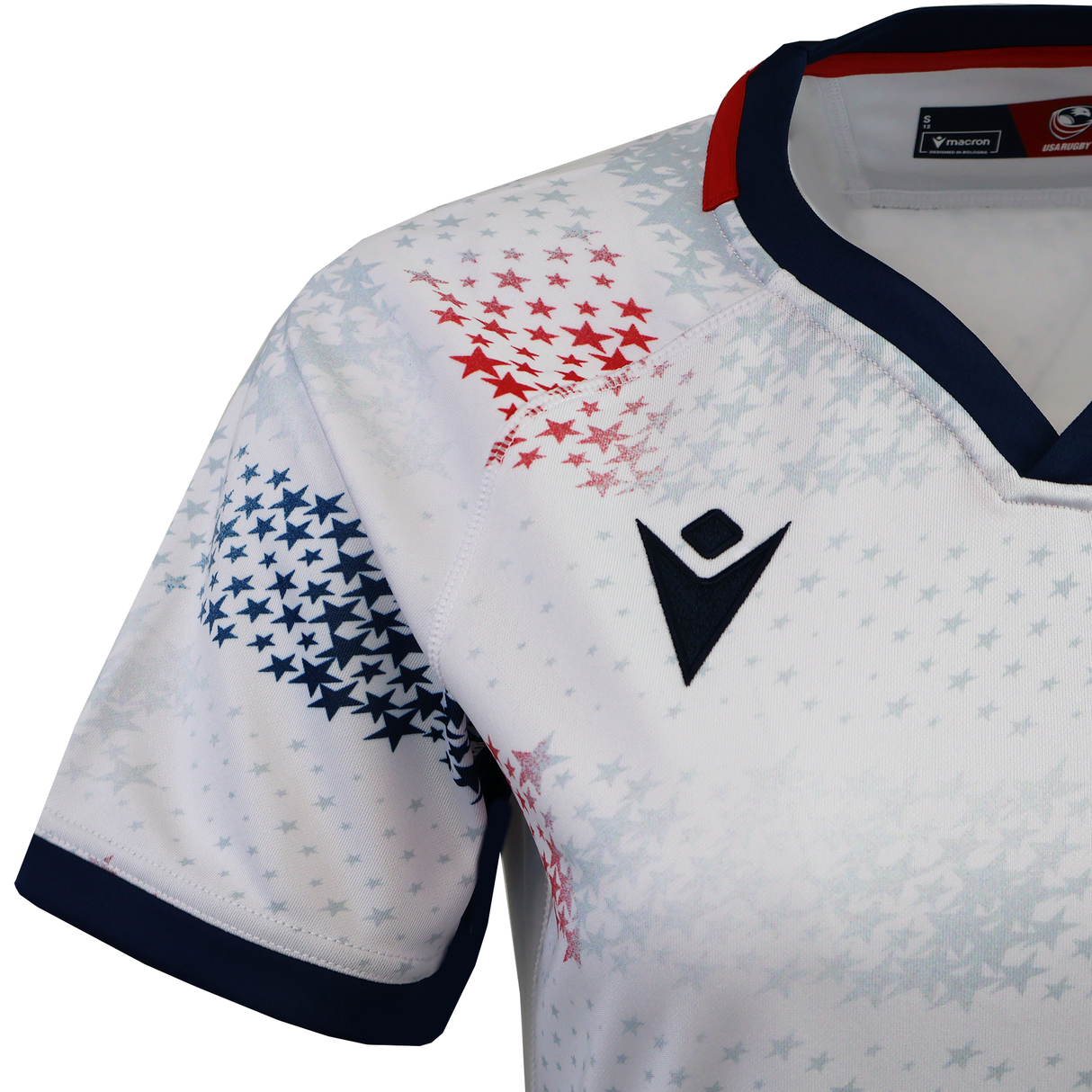 The Women's USA Rugby M25/26 Home Replica Jersey by Macron features a white base with navy blue and red accents, star patterns on the shoulder and chest, and a black logo on the front.