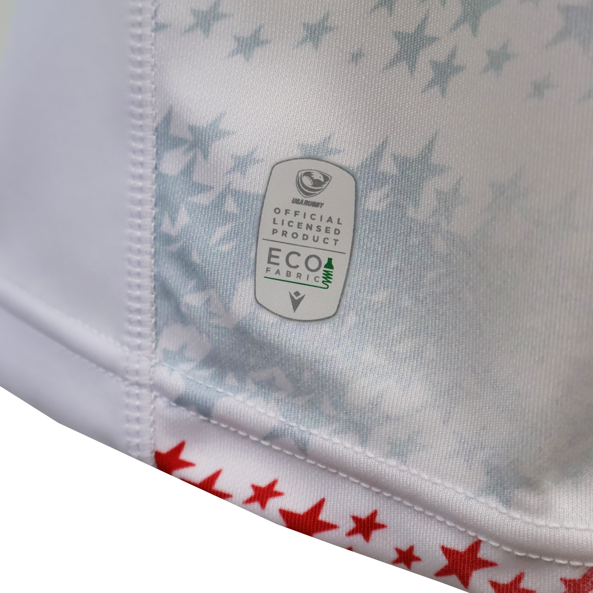 Close-up of the Women's USA Rugby M25/26 Home Replica Jersey by Macron, featuring white fabric with blue star accents, red stars at the hem, and a label showing “Eco Fabric” and “Official Licensed Product.”.