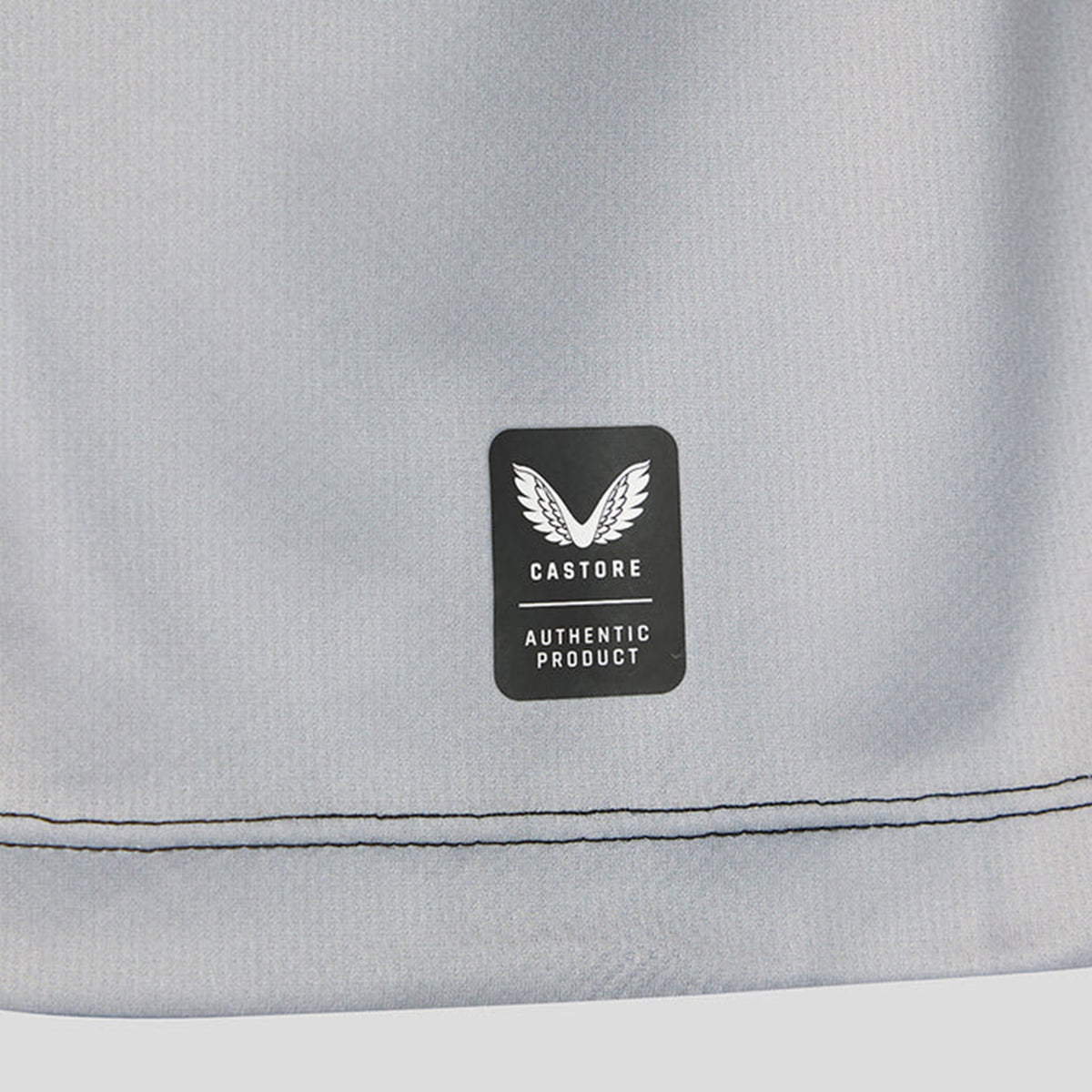 A close-up of gray fabric from the Harlequins 26/27 Replica Home Jersey by Castore, featuring a label with the white winged Castore logo and the text “Castore Authentic Product.” Two lines of stitching can be seen near the fabric’s edge.
