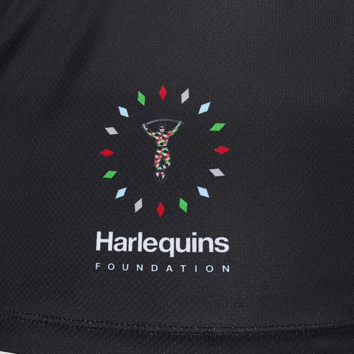 The Harlequins Foundation logo is displayed on black fabric and features a jester-like figure framed by colorful diamond shapes, with the “Harlequins Foundation” text below. This design draws inspiration from the iconic Harlequins 26/27 Replica Third Jersey by Castore and echoes the vibrant style characteristic of Castore’s celebrated rugby shirts.