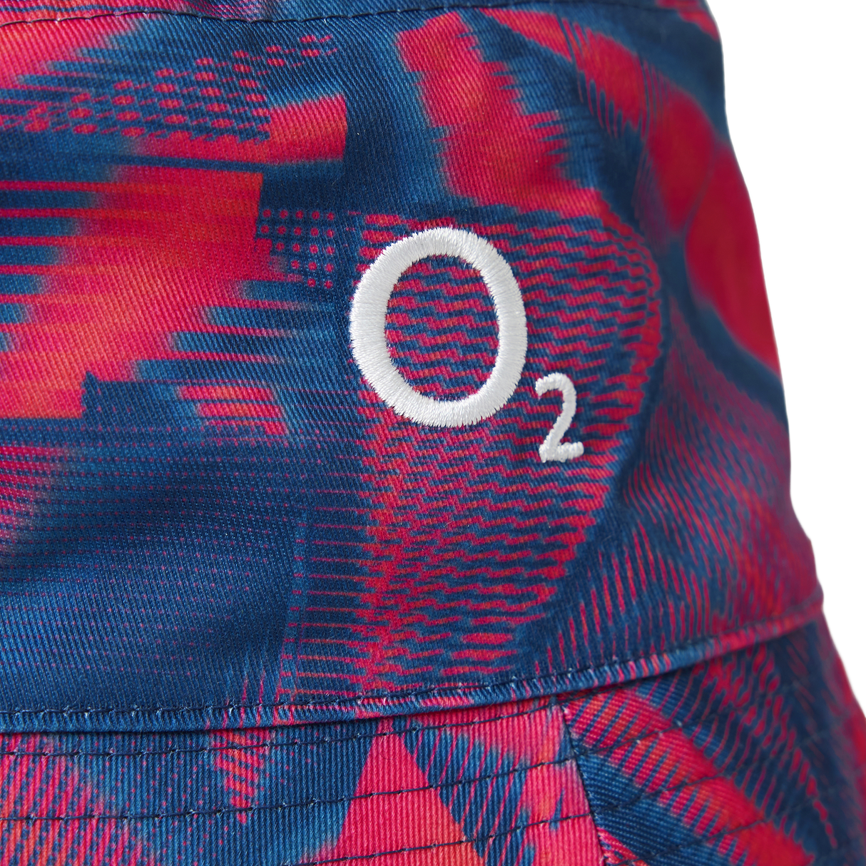 Close-up of a fabric featuring a red and blue geometric pattern with a white “O₂” logo embroidered on it—this detail highlights the premium finish of the Castore England 26/27 Reversible Bucket Cap, designed for lightweight sun protection with modern style.