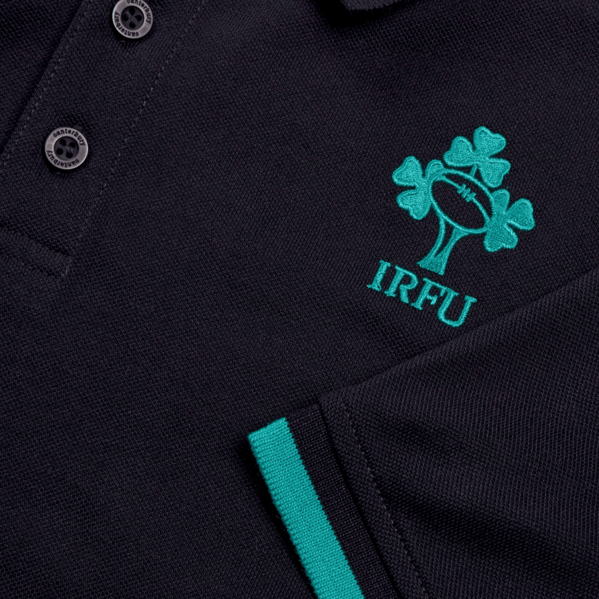 The Ireland 24 Team Polo by Canterbury is a navy cotton shirt featuring embroidered logos, including the IRFU emblem with a green shamrock and rugby ball on the chest. It includes a three-button placket, ribbed collar and cuffs, plus green-striped edge details for added style.