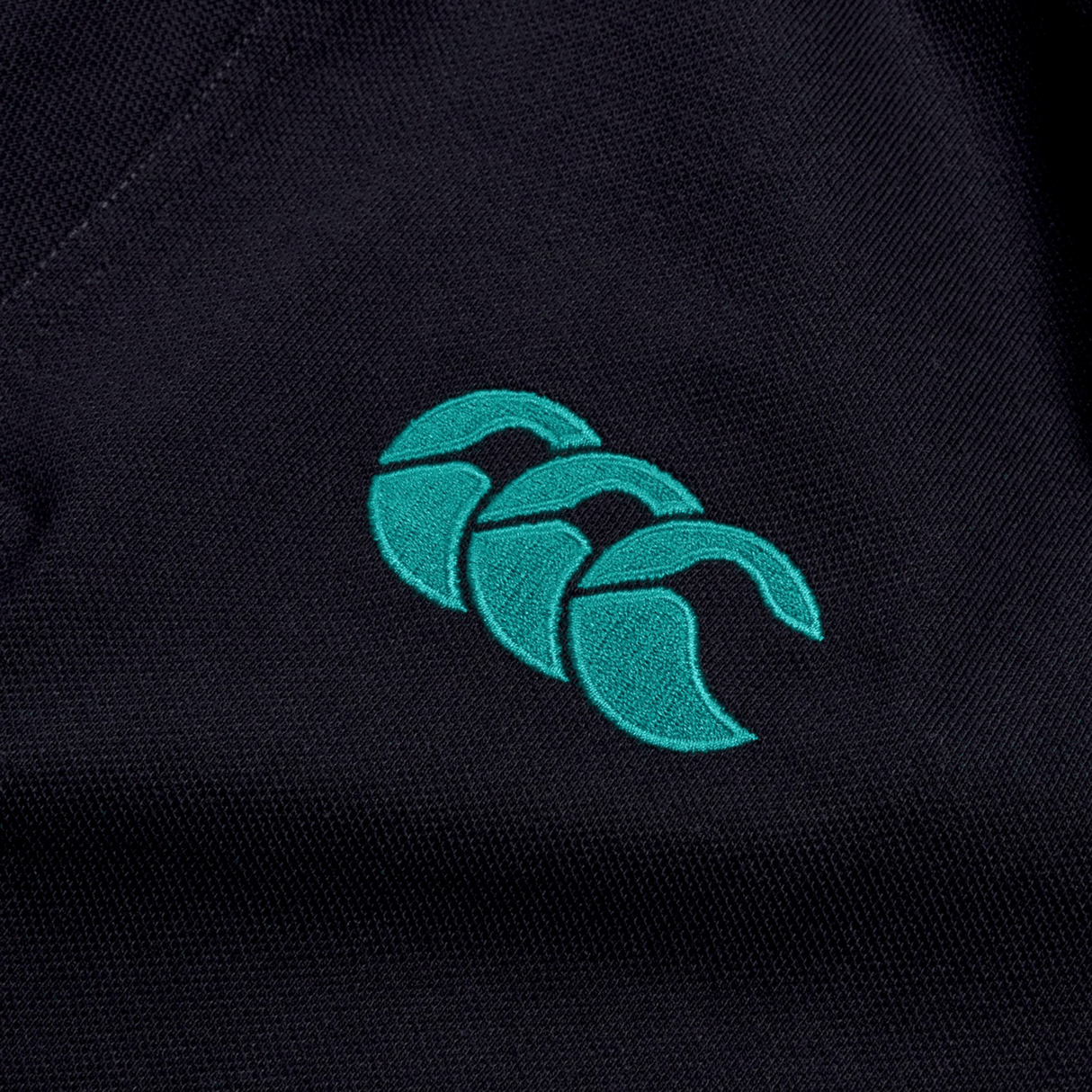 The Ireland 24 Team Polo by Canterbury features a close-up of expertly embroidered teal logo with three overlapping shapes on a dark background, complete with a ribbed collar and cuffs.