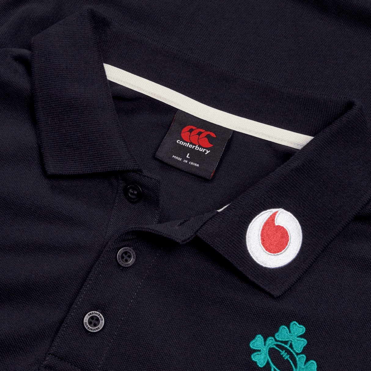 A close-up of the Ireland 24 Team Polo by Canterbury highlights a ribbed collar with a Canterbury tag, featuring a red and white circular logo on the collar and an elegantly embroidered green shamrock below.