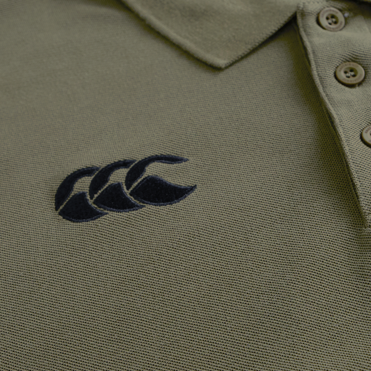 Close-up of the Ireland 25/26 Team Polo by Canterbury in olive green, featuring a collar, three buttons, and a black embroidered logo with three overlapping shapes on the left chest.