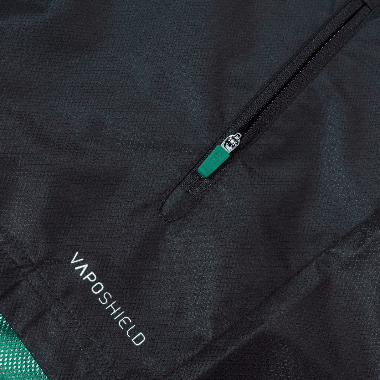 Close-up of a black Ireland 24 Wind Resistant Jacket by Canterbury featuring a zippered pocket with a green pull tab. The "VAPOSHIELD" text is visible near the bottom, highlighting its advanced Vaposhield technology.