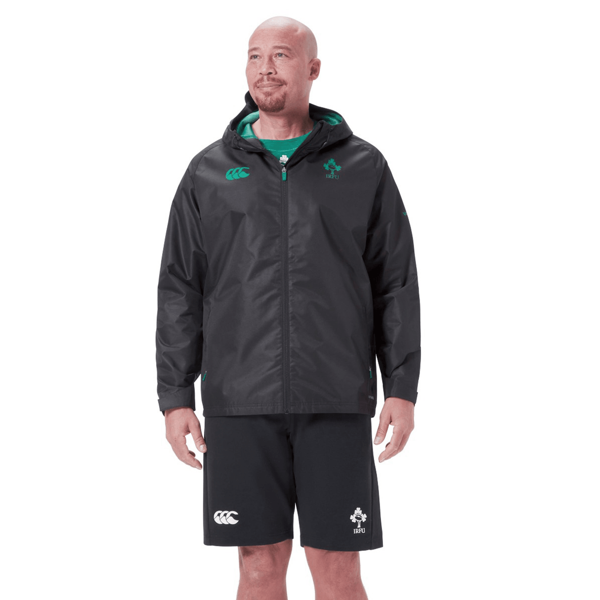 A person with a shaved head is wearing the Ireland 24 Wind Resistant Jacket by Canterbury, which incorporates Vaposhield technology, along with black shorts. Both items feature green accents and logos from the Canterbury brand.