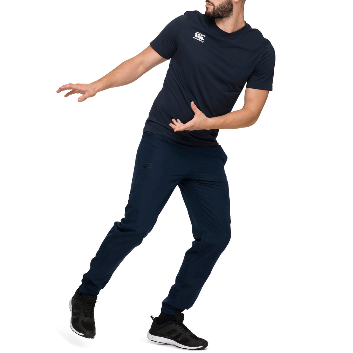 A man in a navy athletic outfit, which includes Canterbury's Team Tonal Taper Leg Cuffed Trackpants, strikes a dynamic pose on one leg against a white background.