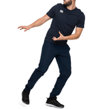 A man in a navy athletic outfit, which includes Canterbury's Team Tonal Taper Leg Cuffed Trackpants, strikes a dynamic pose on one leg against a white background.