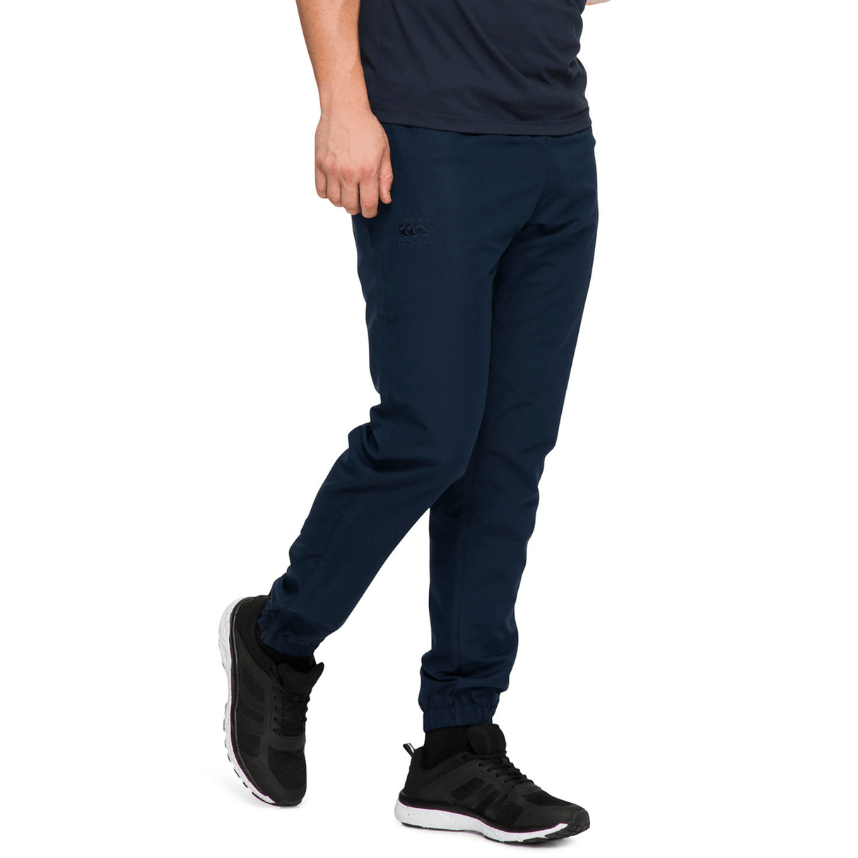 A man in a navy T-shirt and Canterbury Team Tonal Taper Leg Cuffed Trackpant paired with black sneakers stands against a white background.