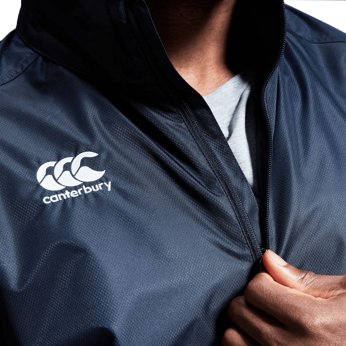 Close-up of a person zipping up a Canterbury Club Vaposhield Full Zip Rain Jacket in navy blue, with the Canterbury logo visible on the left chest. Equipped with Water Resistant VapoShield Technology, this jacket is perfect for rainy days.