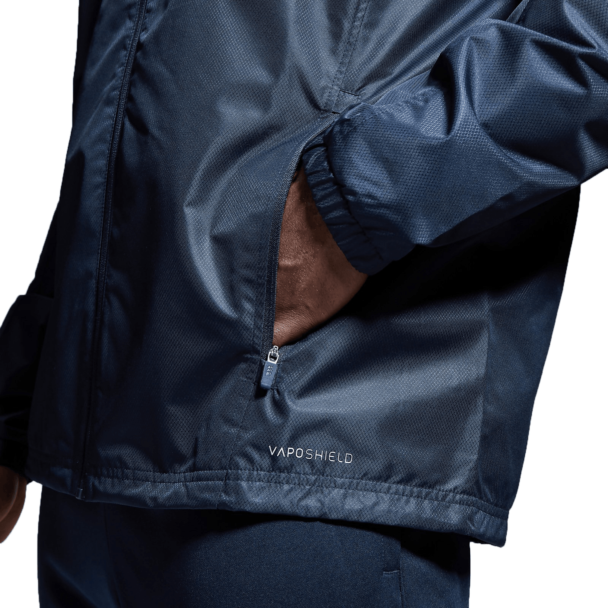 A person wearing a Canterbury Club Vaposhield Full Zip Rain Jacket with water-resistant VapoShield technology has one hand in the zippered pocket. Only the lower half of the jacket and part of the arm are visible.