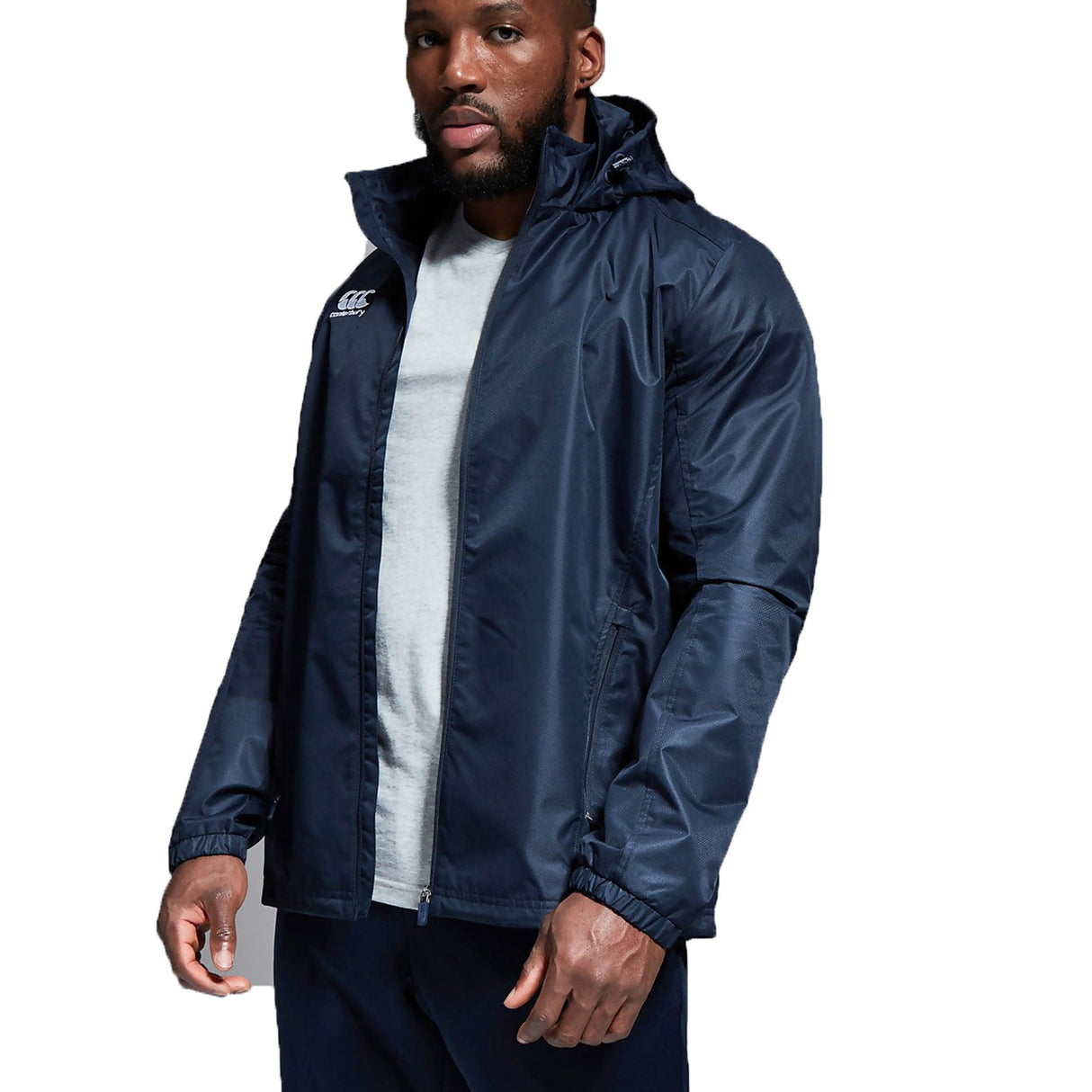 A man wearing a Canterbury Club Vaposhield Full Zip Rain Jacket in navy blue, paired with a light grey shirt and dark pants, poses against a white background.