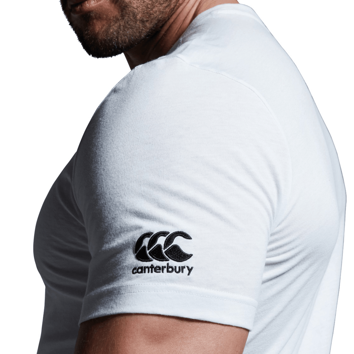 A man is wearing a white Canterbury Florida CCC Flag Club T-shirt by WRS Canterbury, which features the black Canterbury logo and text on the left sleeve. The image highlights his upper torso and arm, with the distinctive CCC Kiwi logo clearly visible against a white background.
