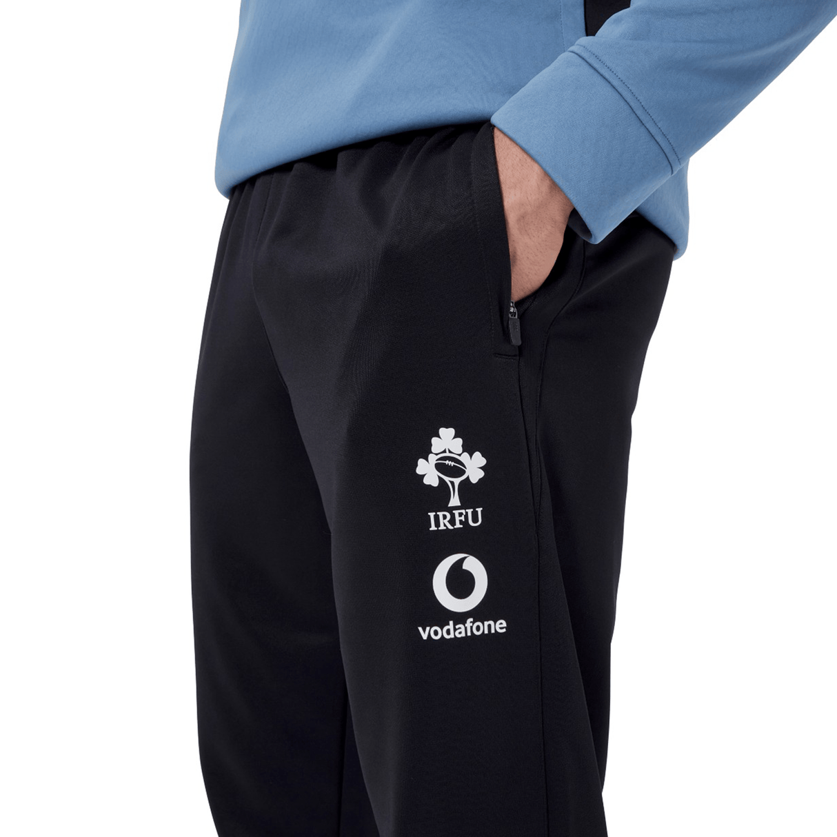 An Irish rugby fan with their hand in the pocket of Canterbury's Ireland 24 Poly Knit Pant, showcasing the IRFU and Vodafone logos, is wearing a long-sleeve blue top equipped with VapoDri technology.