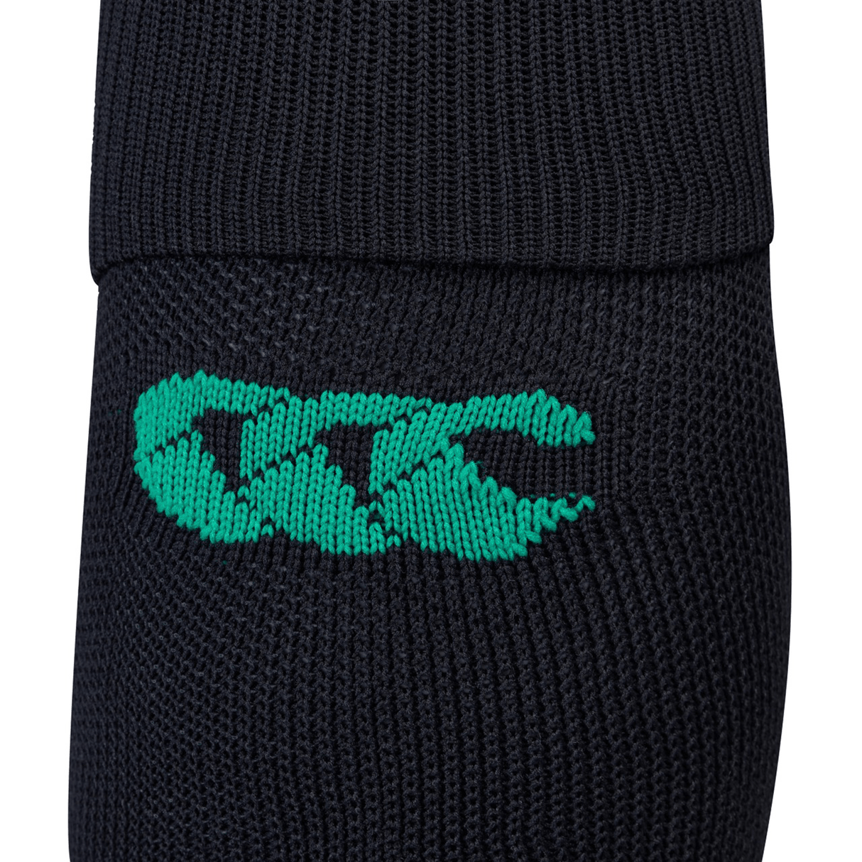 A close-up view of the Ireland 24 Alternate Sock by Canterbury showcases its black knitted fabric adorned with a green logo consisting of three stylized overlapping letters. The sock includes ankle and arch support and features fast-drying performance yarn.