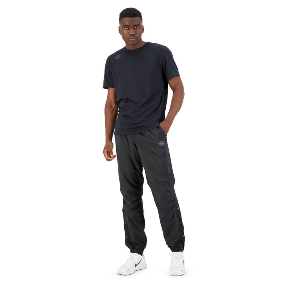 A young man wearing a casual dark outfit with a Canterbury Tempo logo on his shirt and Canterbury Vaposhield Dome Pants, standing confidently against a plain white background.