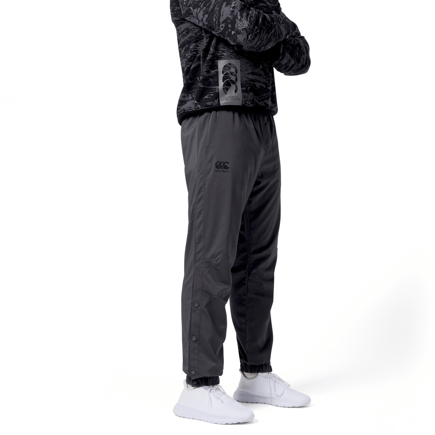 A person stands confidently with arms crossed against a white background, wearing a dark camouflage sweater and Canterbury Tempo Vaposhield Dome Pants with an elastic waistband, paired with white sneakers.