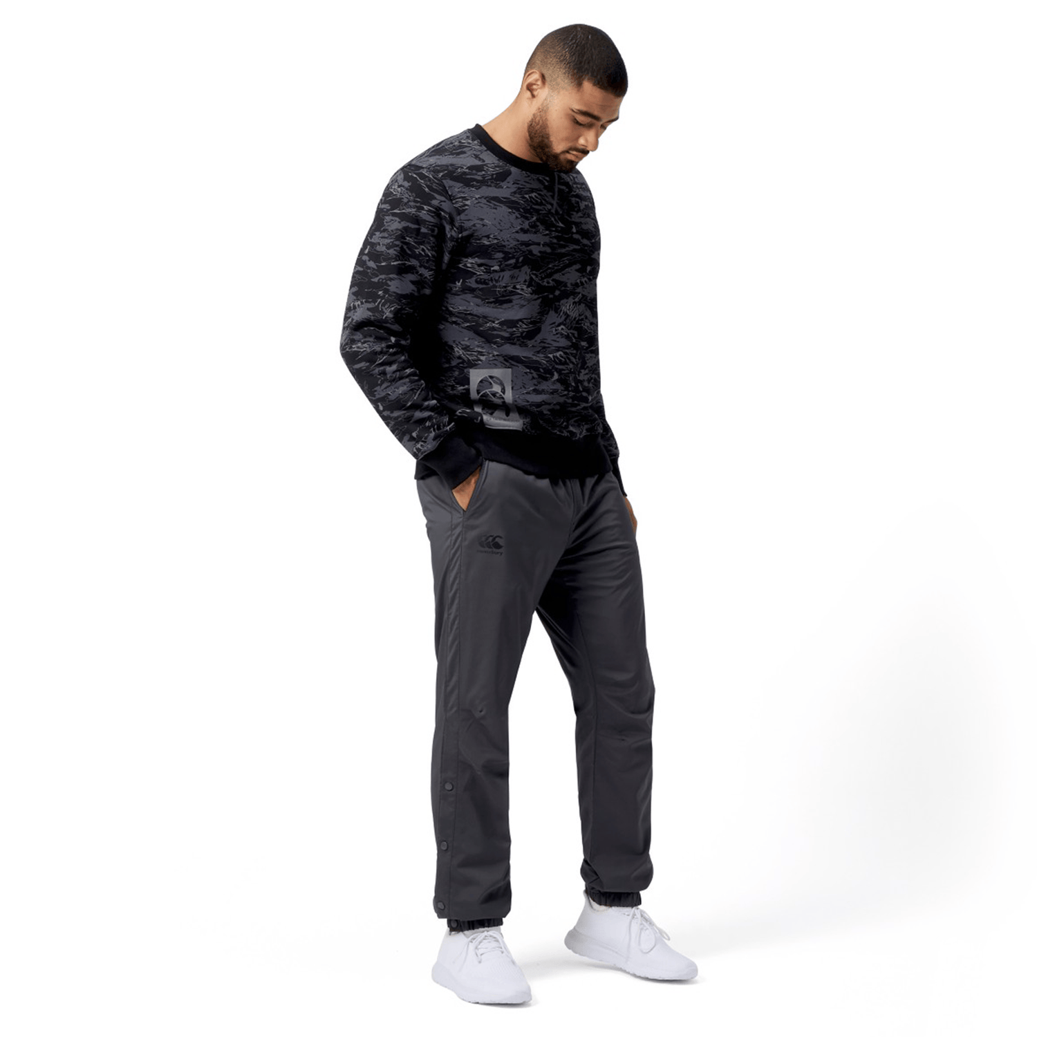 A man in a camo-patterned sweatshirt, Canterbury Tempo Vaposhield Dome Pants, and white sneakers stands against a white background, looking down with his hands in his pockets.
