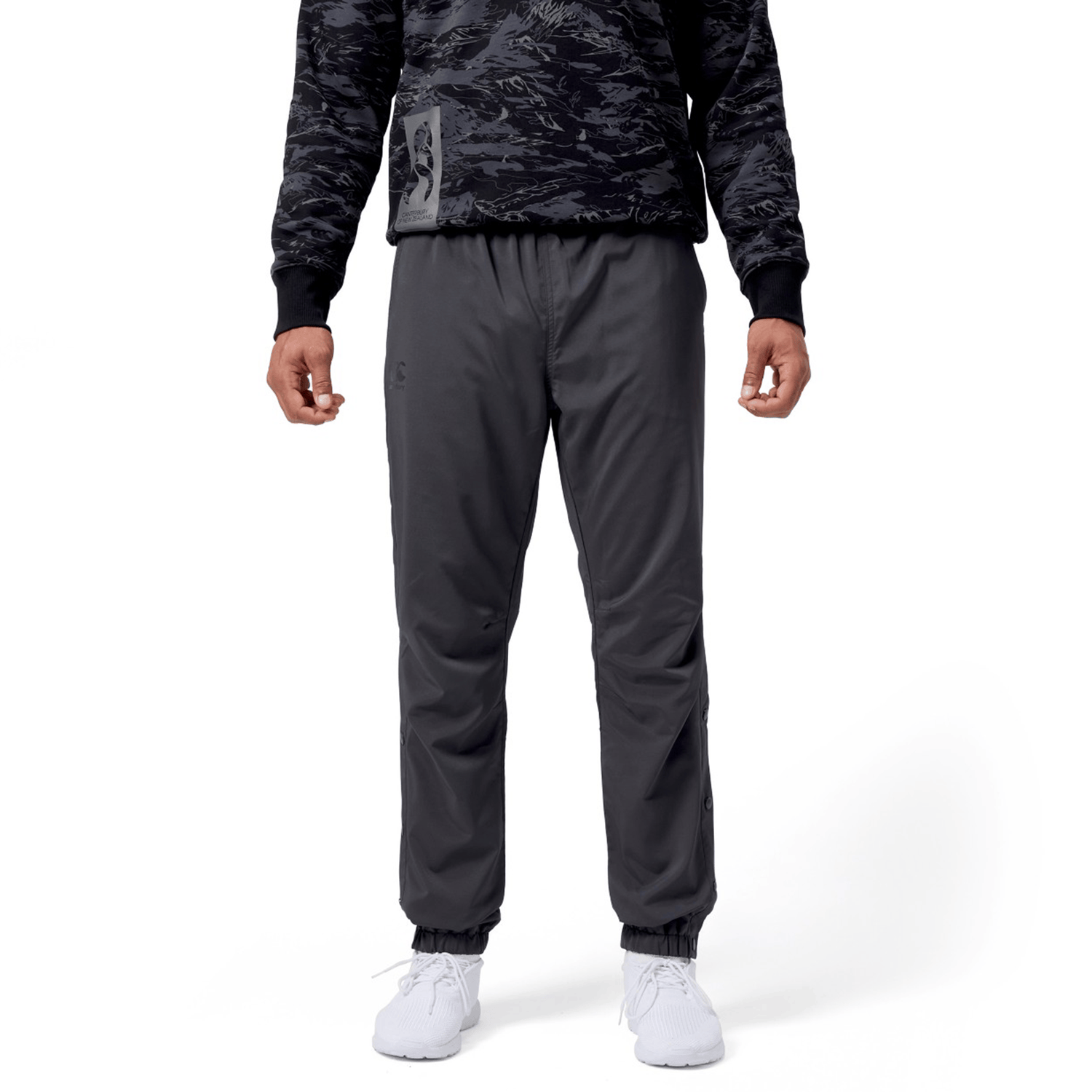Against a white backdrop, a person wears a dark camo hoodie, Canterbury Tempo Vaposhield Dome Pants with an elastic waistband, and white sneakers.