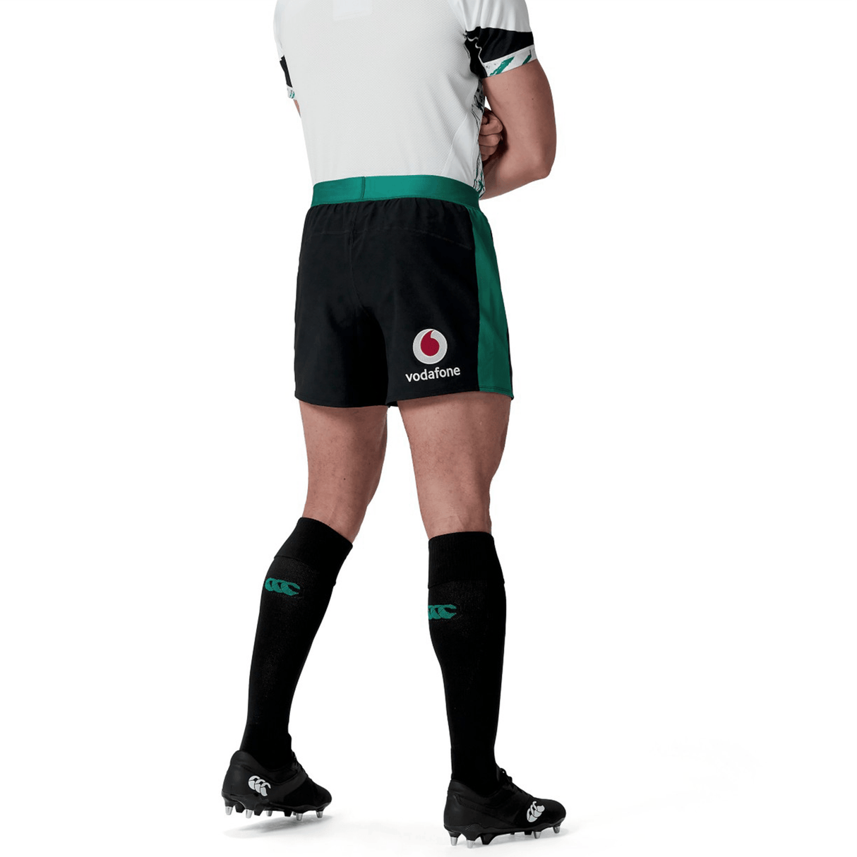 A person wearing a white shirt, Ireland 24 Alternate Match Short by Canterbury featuring a Vodafone logo, black socks, and black cleats stands with hands behind their back.