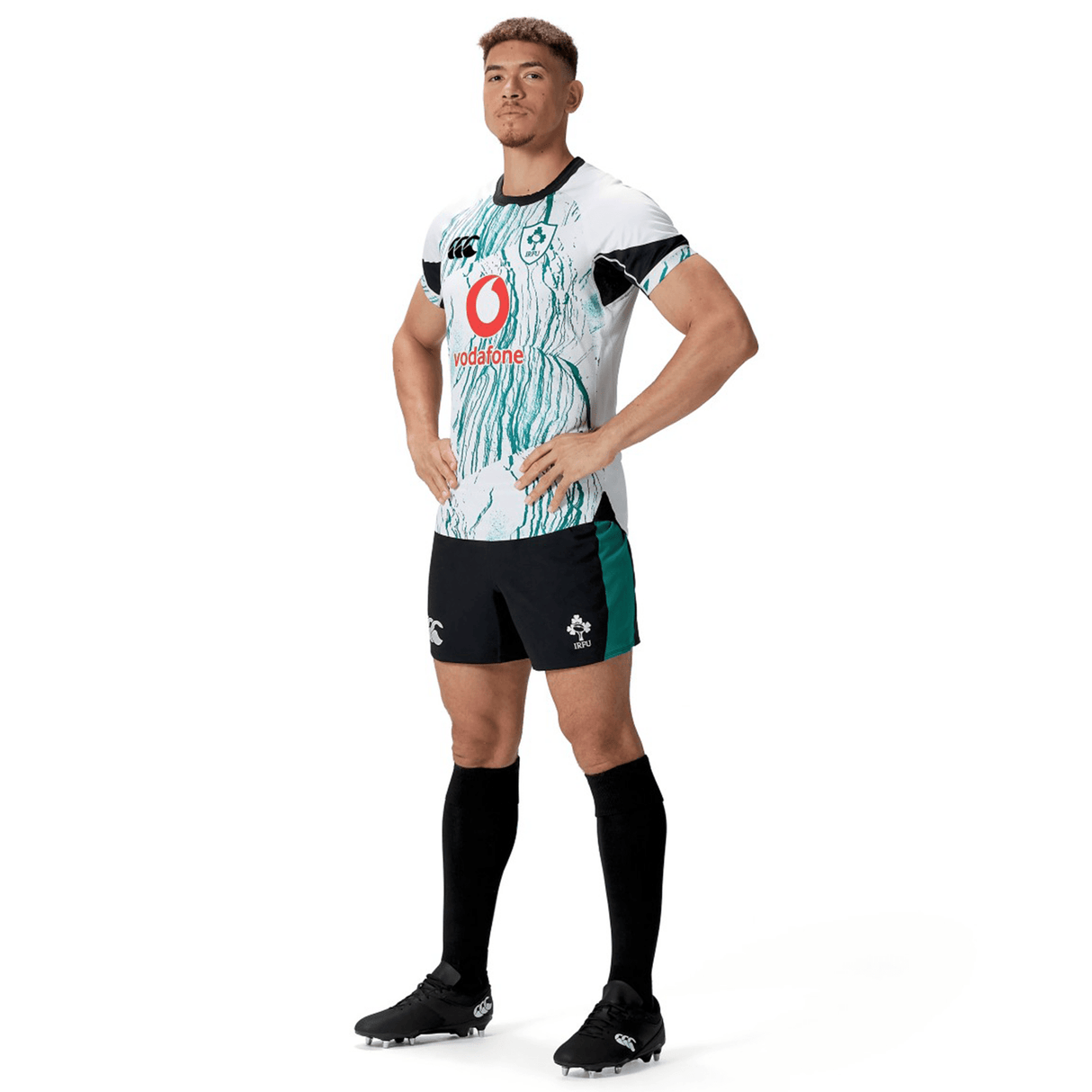 A rugby player is posing confidently in a white and green jersey, Canterbury's Ireland 24 Alternate Match Short made from moisture-wicking fabric, and black socks. He has short hair and stands with his hands on his hips.