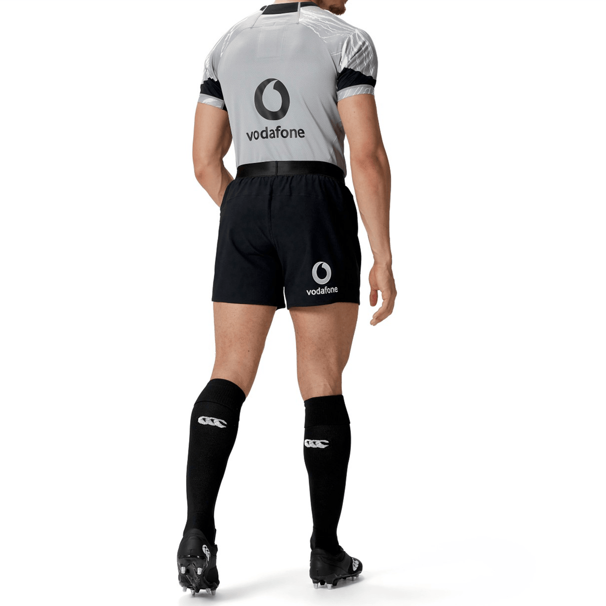 A person is standing with their back to the camera, clad in a black and gray sports uniform from Canterbury featuring the "vodafone" logo on the shirt. They are also wearing the Ireland 24 Training Short by Canterbury, paired with black knee-high socks and cleats.