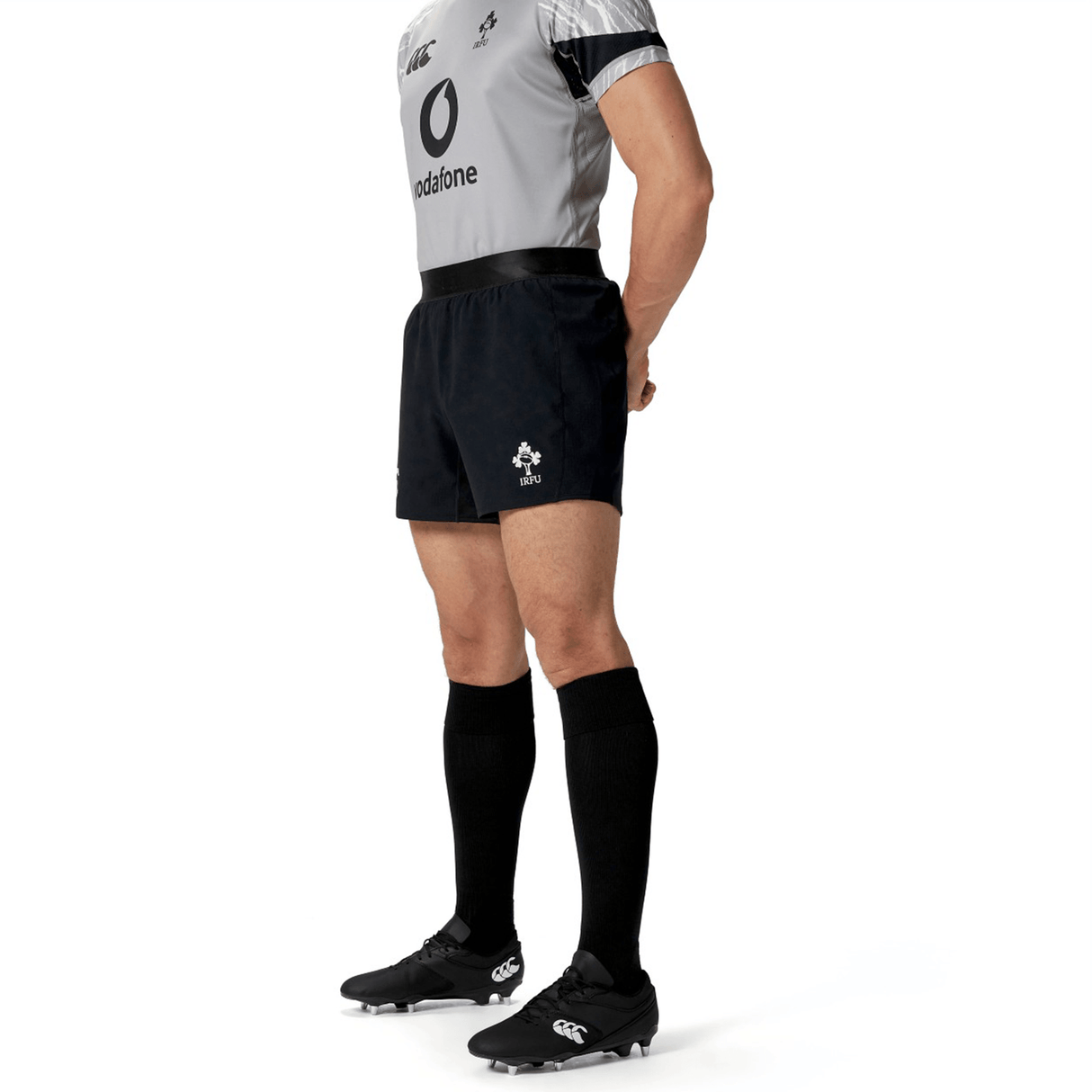 A person in rugby training apparel stands with hands behind back. The uniform includes a grey jersey, Canterbury's Ireland 24 Training Short in black, black socks, and cleats.