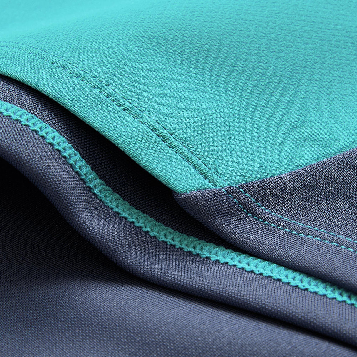 Close-up of two pieces of fabric, one teal and one dark blue, sewn together with visible stitching and seams, featuring details characteristic of the Ireland 26 Tech Drill Top by Canterbury.
