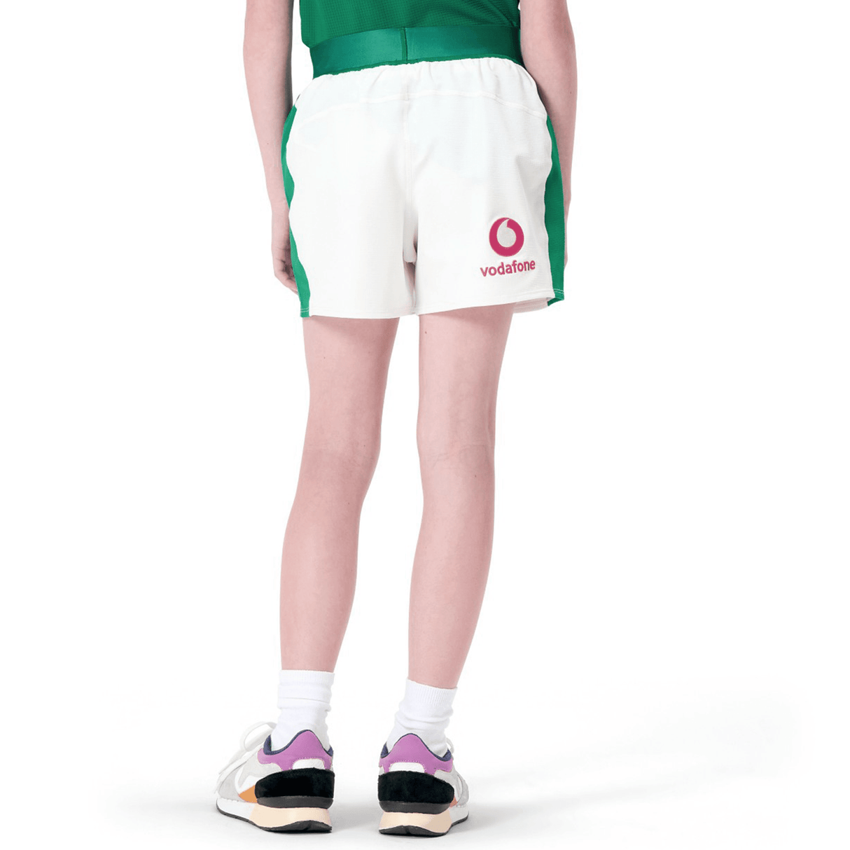 A person stands in a green shirt paired with Junior Ireland 24 Home Match Shorts by Canterbury, crafted from moisture-wicking fabric and featuring the "Vodafone" logo. Wearing sneakers with pink and black accents, they face away.