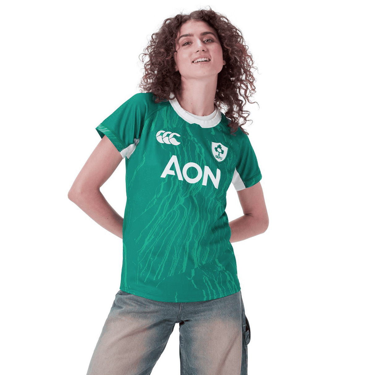 A person is wearing a green Women's Ireland WIRFU Home Pro Jersey by Canterbury, featuring VapoDri technology and the "AON" logo. They pair it with light jeans against a white background, proudly displaying the official IRFU crest.