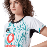 A person with curly hair wears the Women's Ireland 24 Alternate Pro Jersey by Canterbury, a white and green jersey showcasing the iconic IRFU Crest.