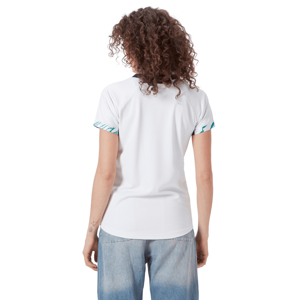 A person with curly hair faces away from the camera, wearing a white T-shirt with patterned sleeves, similar to the Women's Ireland 24 Alternate Pro Jersey by Canterbury, and blue jeans.