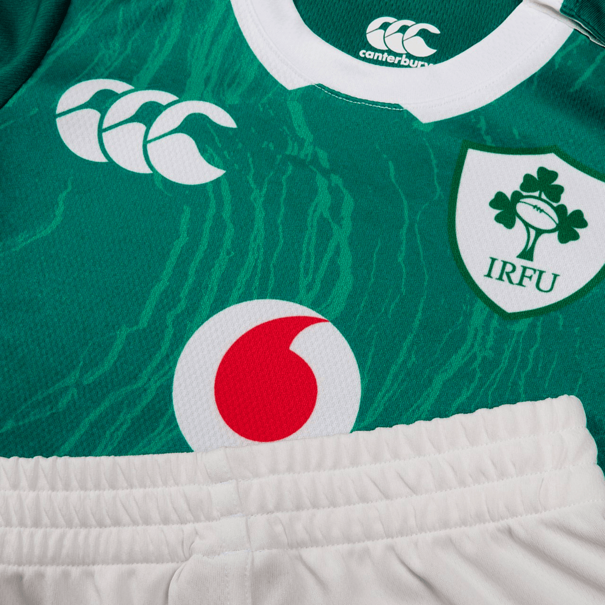 Close-up of a green Infant Ireland 24 Home Pack rugby jersey by Canterbury, featuring the IRFU and Canterbury logos, with a partially visible red and white sponsor logo, paired with white shorts.