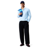A man faces forward against a white background, wearing the Canterbury Solid Dye Rugby Jersey by Canterbury in blue, dark trousers, a navy cap, loafers, and holding a folded striped towel over his left shoulder.