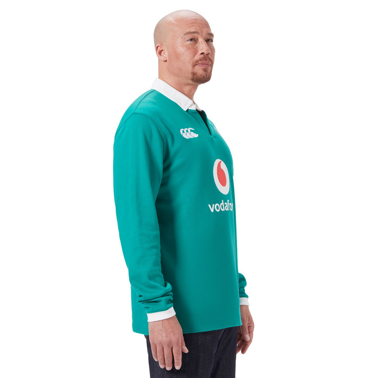 A bald man stands in profile wearing the Ireland 25/26 Long Sleeve Classic Home Jersey by Canterbury, featuring a red and white logo on the front, paired with dark jeans.