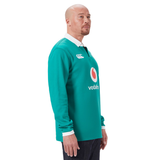 A bald man stands in profile wearing the Ireland 25/26 Long Sleeve Classic Home Jersey by Canterbury, featuring a red and white logo on the front, paired with dark jeans.
