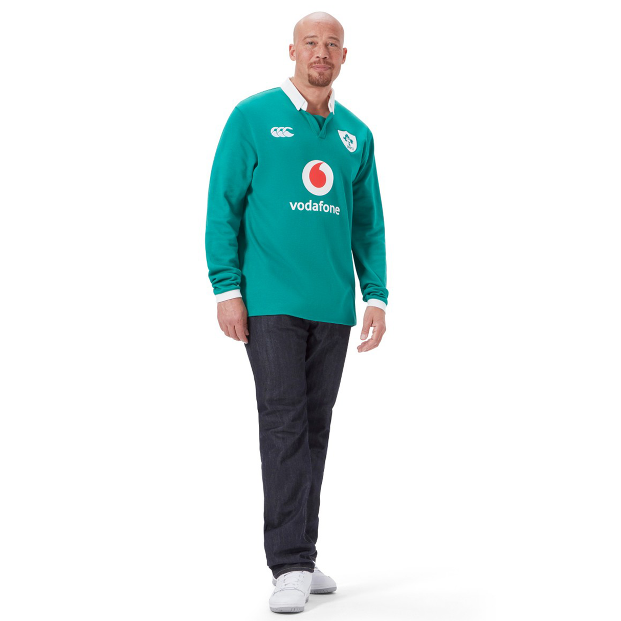 A man in the Ireland 25/26 Long Sleeve Classic Home Jersey by Canterbury, featuring logos, stands against a white background with dark jeans and white shoes.