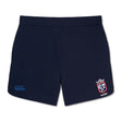 Introducing the Claremont Colleges Elite Woven Short by Canterbury: a pair of navy blue rugby shorts featuring an EMB Canterbury logo on the left leg and a white and red crest on the right leg, perfect for elevating your training experience.