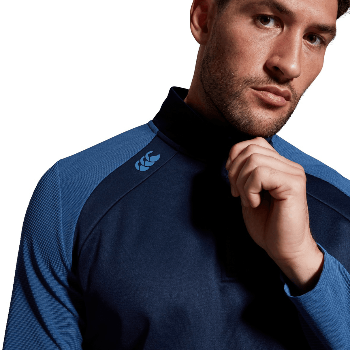 Man in a Canterbury Elite 1/4 Zip blue sports top with a high neck, posing with his hand on his chin, looking thoughtfully towards the camera.