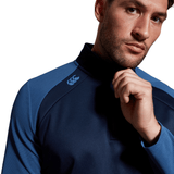 Man in a Canterbury Elite 1/4 Zip blue sports top with a high neck, posing with his hand on his chin, looking thoughtfully towards the camera.