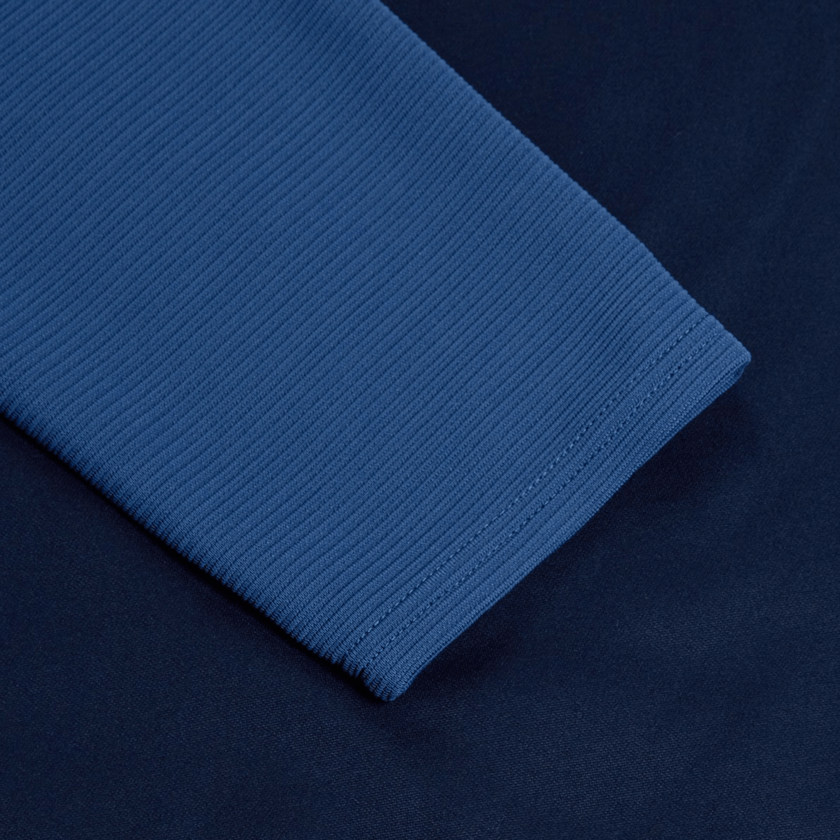 Close-up of a Canterbury Elite 1/4 Zip Top in blue performance fabric with ribbed texture on a darker blue background, highlighting the detailed stitch work at the edge.