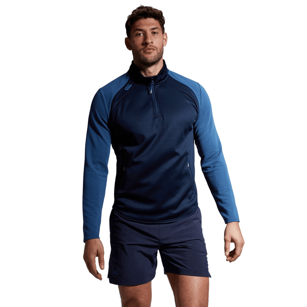 A man in a blue Canterbury Elite 1/4 Zip Top athletic top and black shorts standing confidently against a plain background.