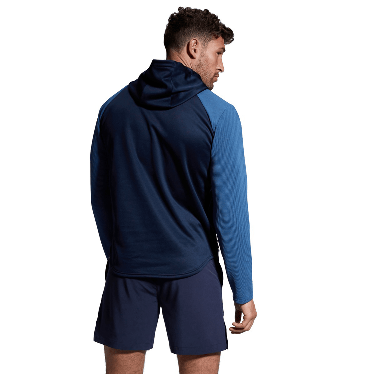 A man dressed in a blue Canterbury Elite Training Hoodie, crafted from premium fabrics, paired with navy shorts, standing with his back turned and looking to the side against a white background.