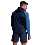 A man dressed in a blue Canterbury Elite Training Hoodie, crafted from premium fabrics, paired with navy shorts, standing with his back turned and looking to the side against a white background.