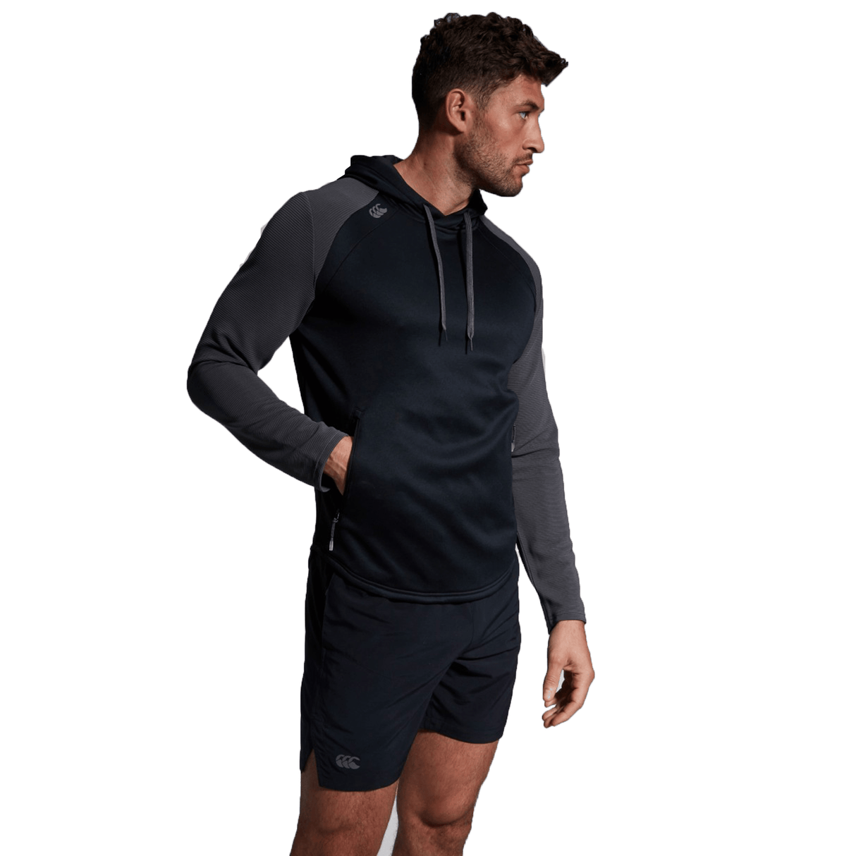 A man in a dark navy and gray Canterbury Elite Training Hoodie made from premium fabrics, paired with black shorts, stands with his left hand in his pocket, looking to the side.