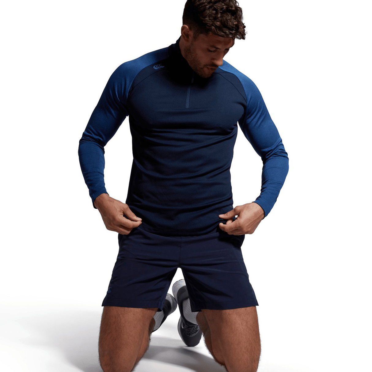A man is crouching down in front of a blue shirt and shorts with the Canterbury Elite First Layer, showcasing VapoDri technology.