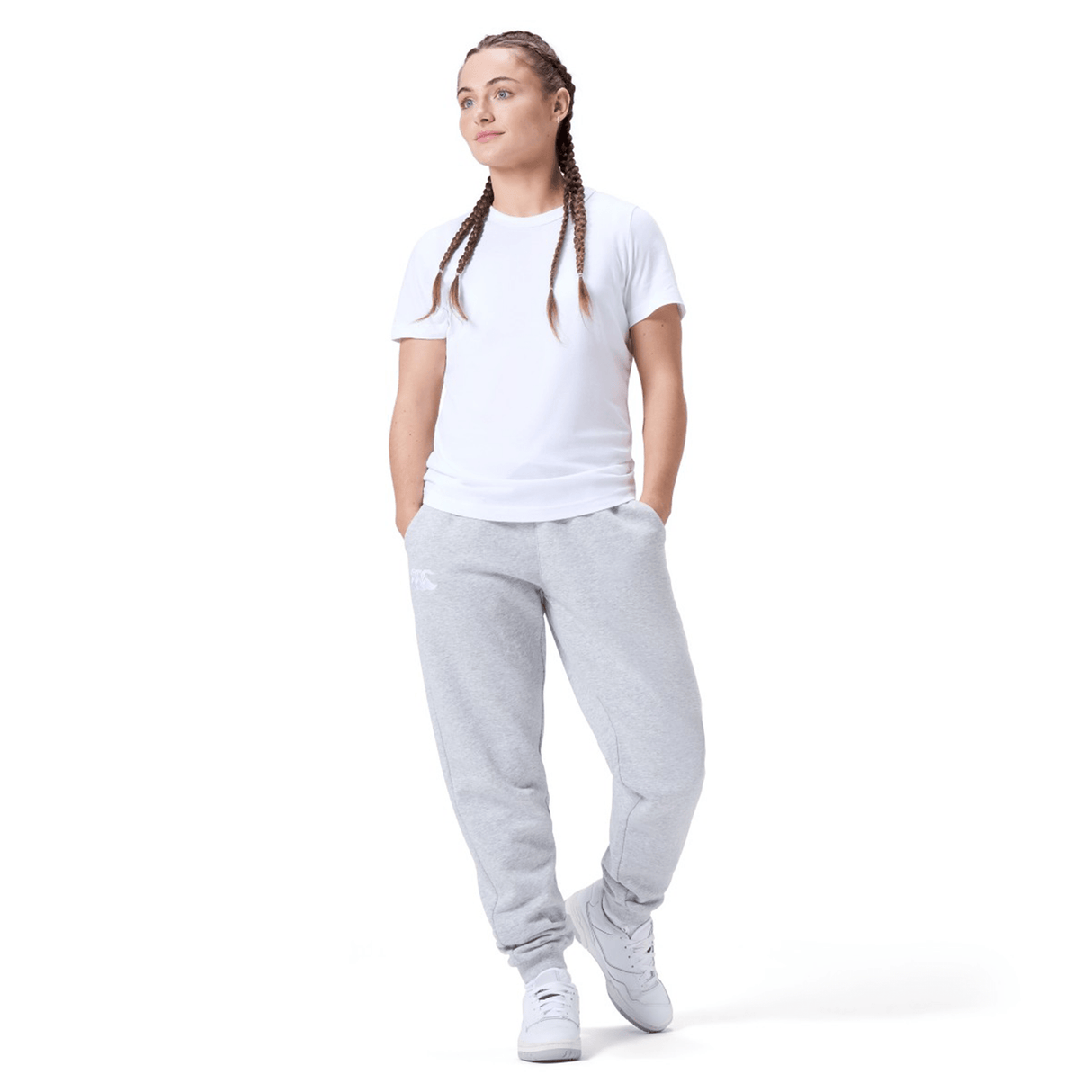 A person in a white t-shirt, Canterbury Women's Leisure Sweatpant by Canterbury, and white sneakers stands with hands in pockets, looking slightly to the side. Their hair is styled in braids, complementing the casual cotton blend fleece outfit perfectly.